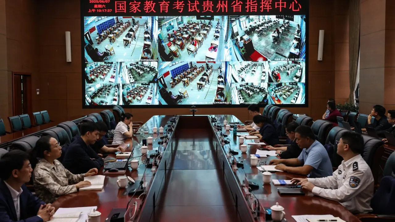 A meeting room with a long table where officials review multiple surveillance camera feeds on a large screen, displaying students taking exams in various locations. Participants are engaged with documents and electronic devices.